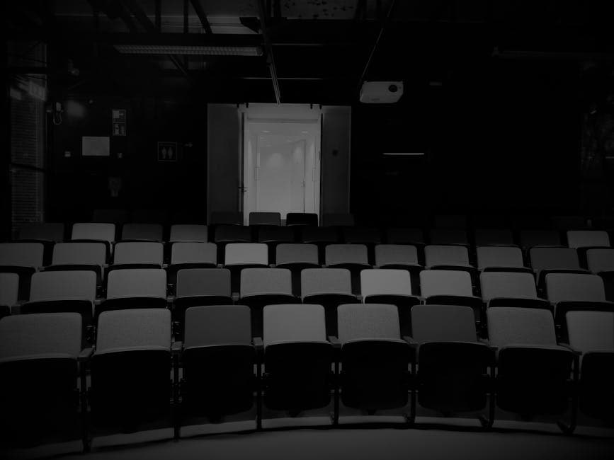 A dimly lit empty auditorium with rows of seats and a bright doorway at the back, captured in monochrome.