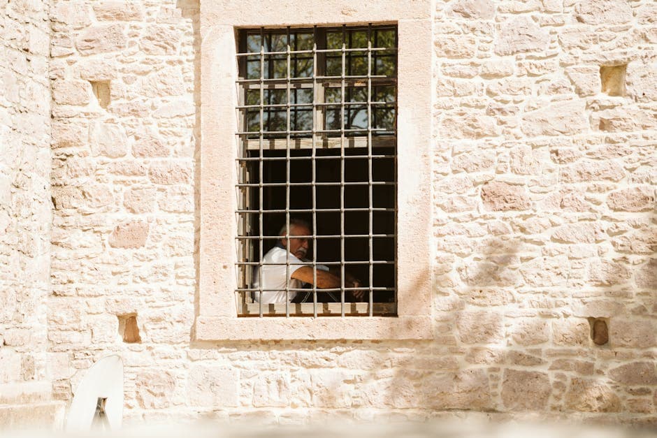An elderly man is seen sitting inside a building, framed by a barred window of a stone wall.