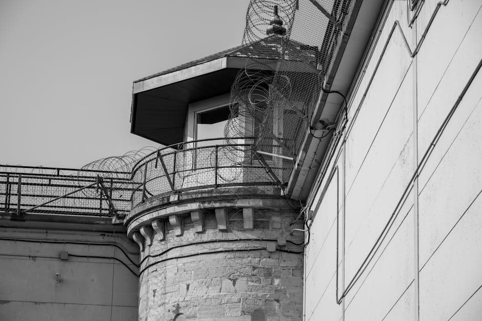 Black and white image of a prison tower secured with barbed wire fencing.