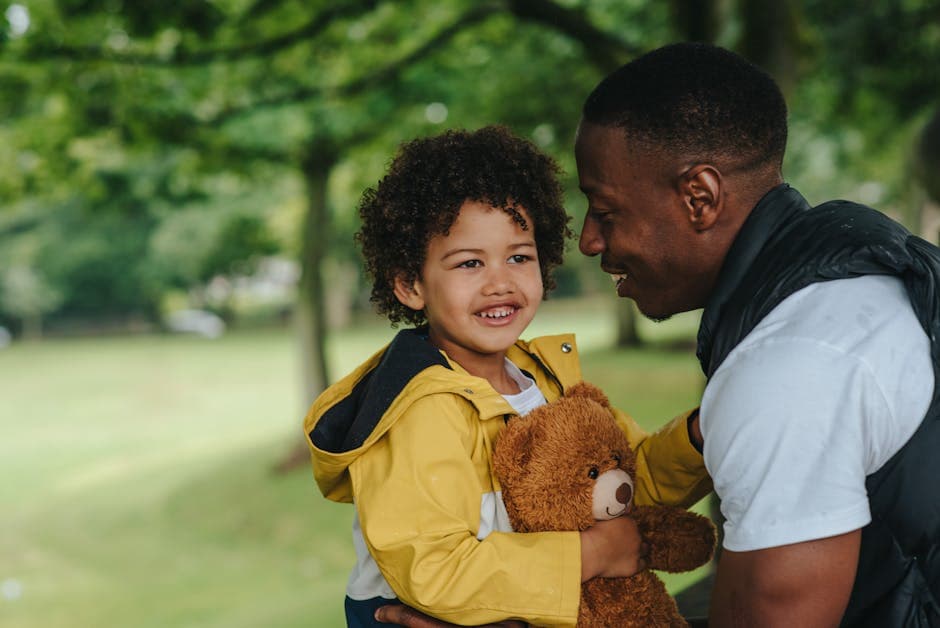 Happy little boy carrying toy bear and looking away while spending free time together with African American father in park