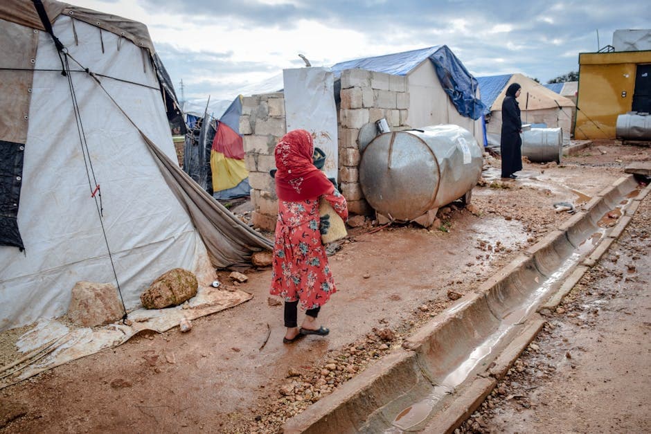 Full body of poor women in hijabs and headscarves near weathered buildings and camps in village under cloudy sky