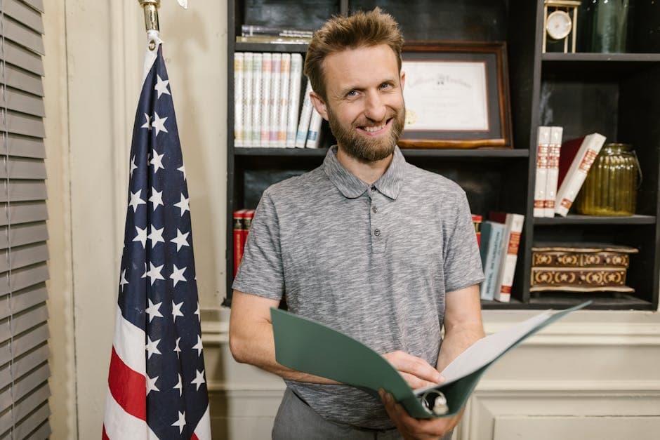 Smiling man in an office holding a folder with an American flag nearby.