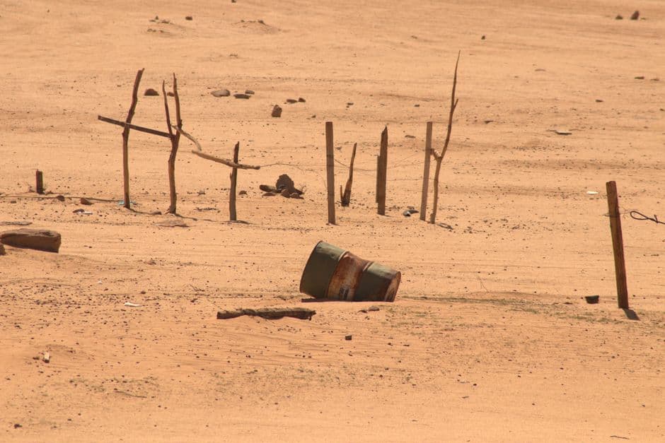 Desert landscape featuring a rusty barrel amid sand, sticks, and sparse vegetation.