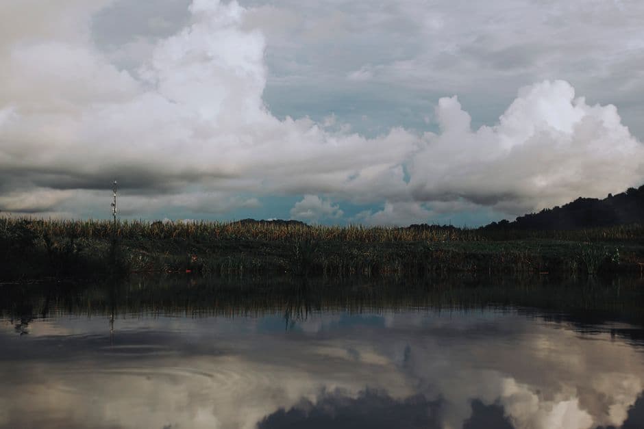 A calm scene with clouds reflecting on a tranquil body of water, surrounded by lush greenery.