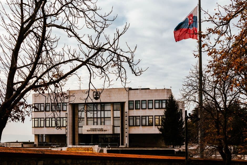 A view of the Slovak Parliament building surrounded by autumn trees with the national flag waving under an overcast sky.