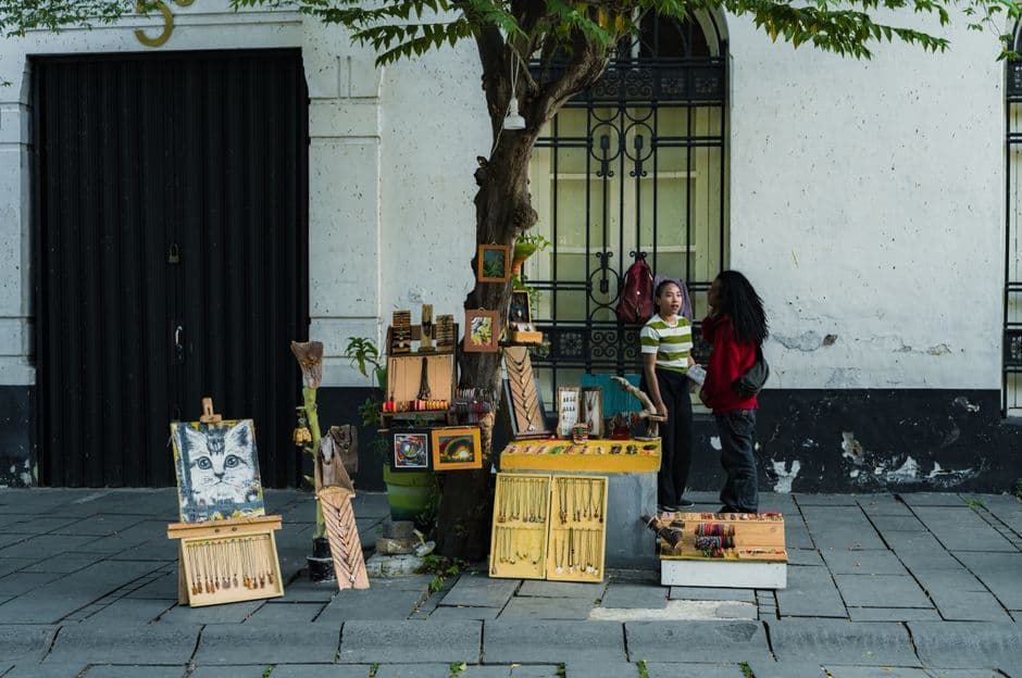 Two people converse at a street vendor stall displaying art and souvenirs on a city sidewalk.