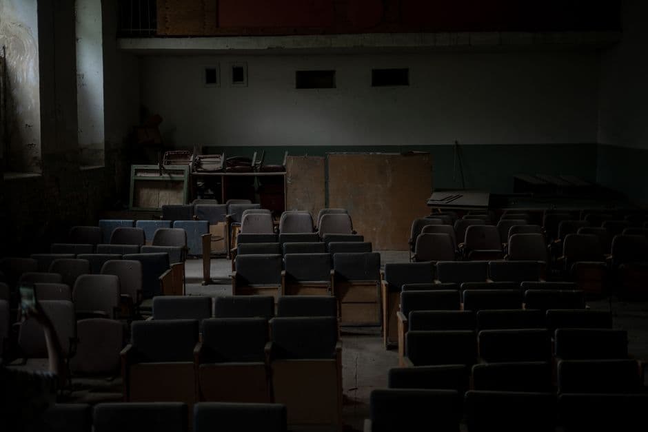 Dimly lit abandoned auditorium with rows of empty chairs and scattered furniture.