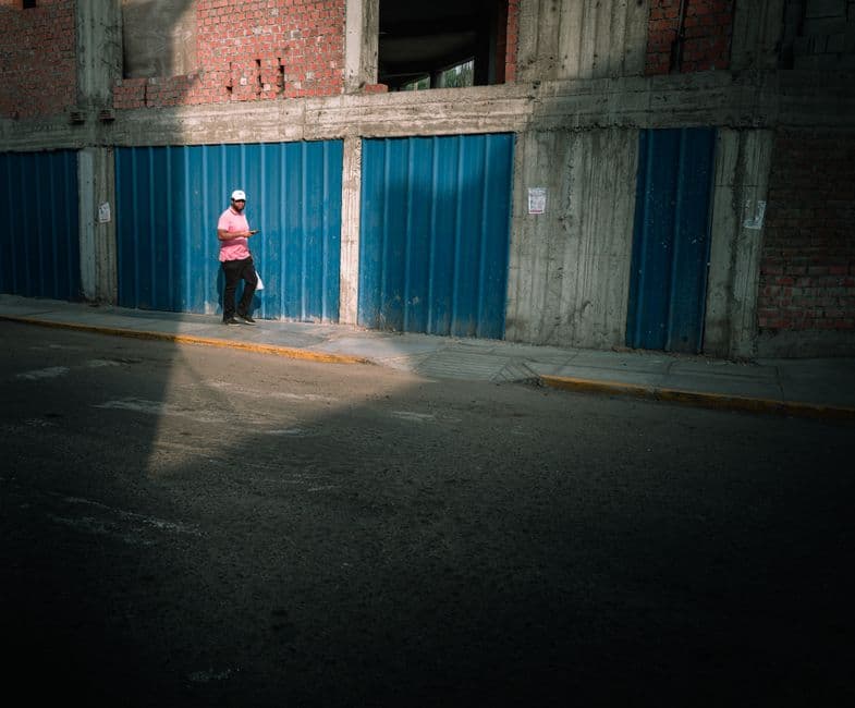 A man walks along a shadowed street beside a building with blue metal panels.