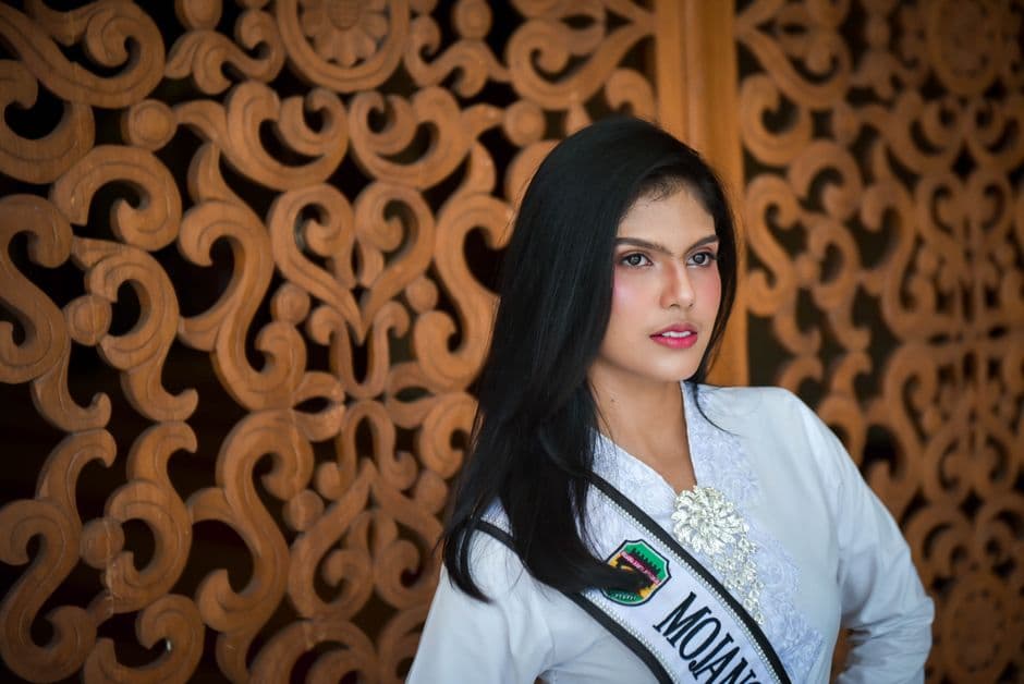 Portrait of an elegant woman in traditional Indonesian attire with intricate wooden background.