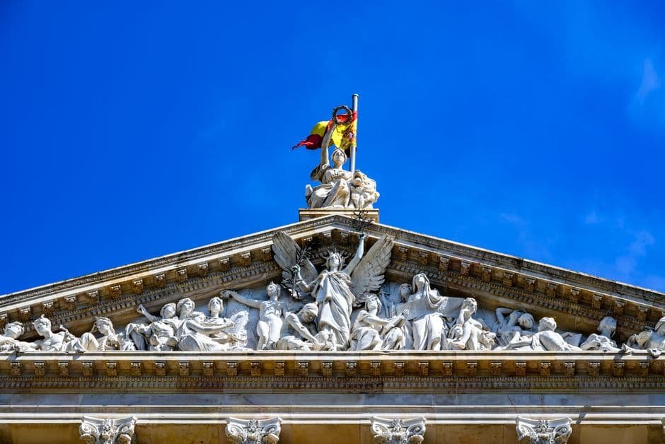 Historic Parthenon-style architecture adorned with sculptures and Spanish flag, captured in vibrant sunlight.