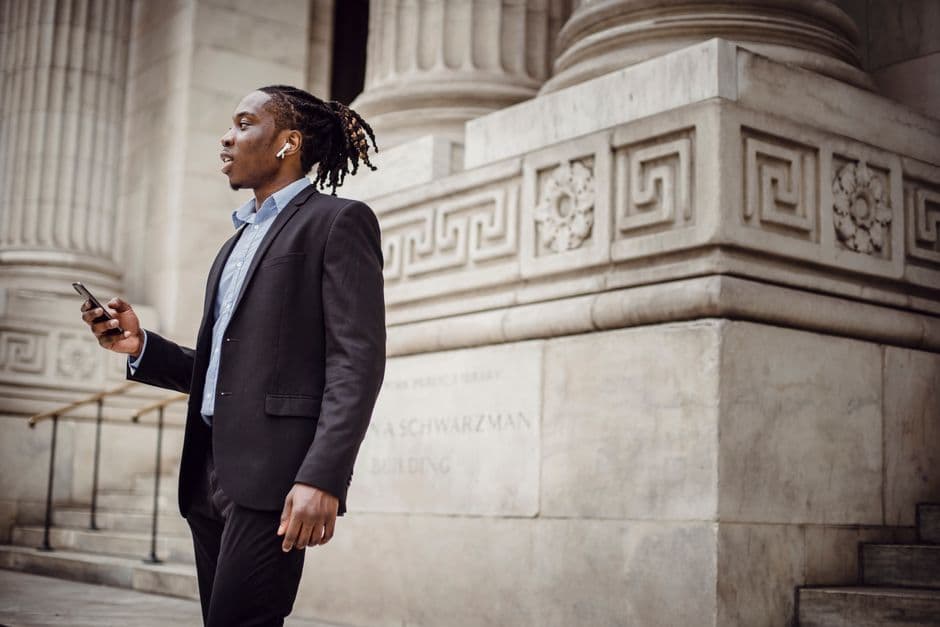 Side view of positive African American man in formal suit standing against historic building and talking on mobile phone using wireless earphones