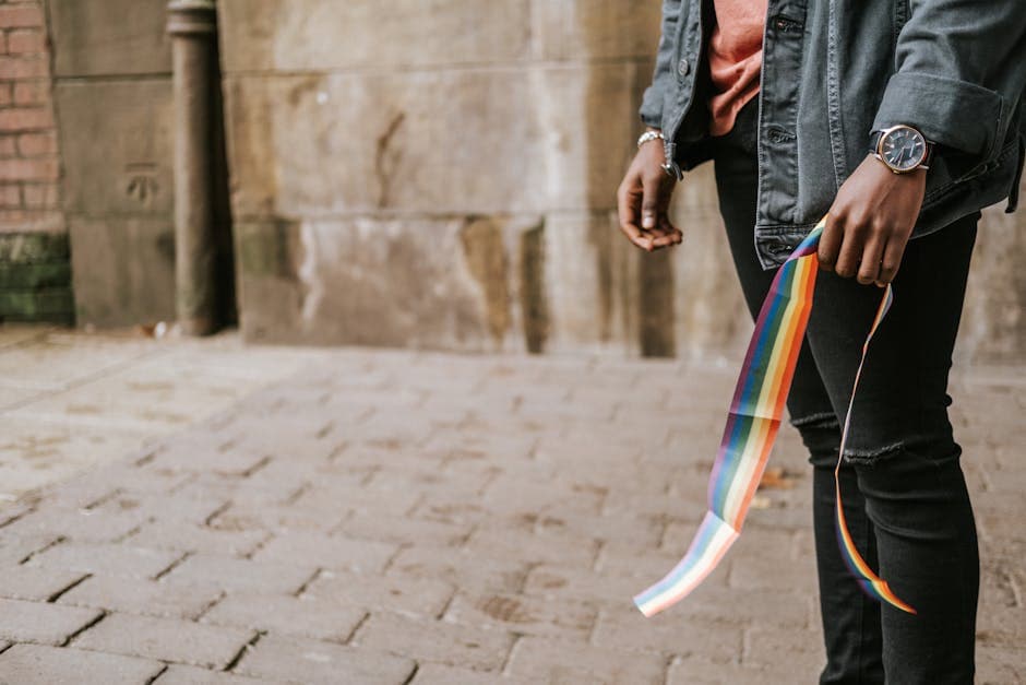 Crop anonymous African American male in casual clothes standing on street sidewalk with colorful rainbow ribbon in hand