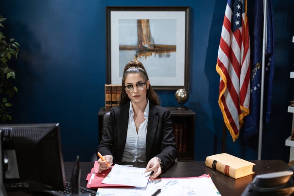 Focused woman in black blazer working on documents at desk with American flag in the background.