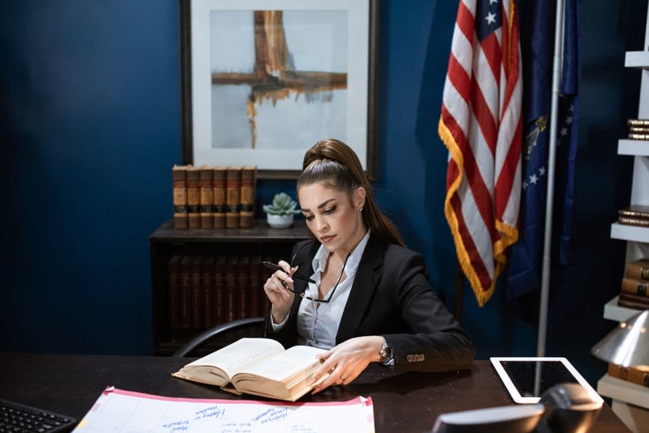 Woman in black blazer reading a book at an office desk, displaying a serious and focused demeanor.