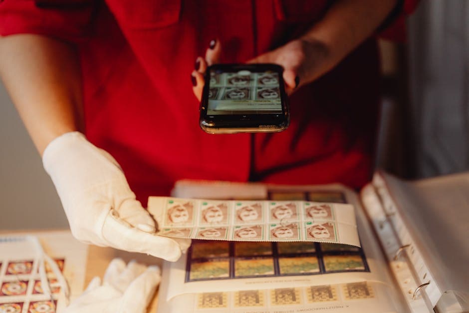 A person in gloves photographs a stamp collection using a smartphone for digital archiving.