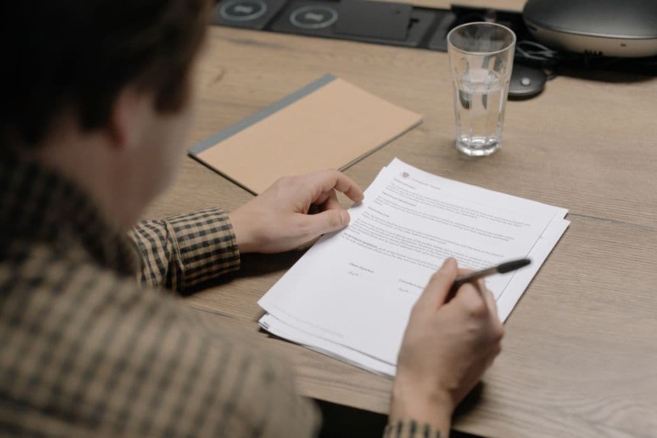 A businessman is reviewing a contract while holding a pen at a desk with a glass of water nearby.
