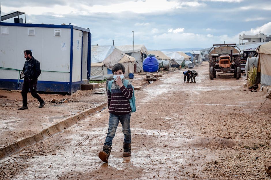 Full body of ethnic boy in protective mask strolling on dirty path along shabby tents in poor district of countryside during pandemic