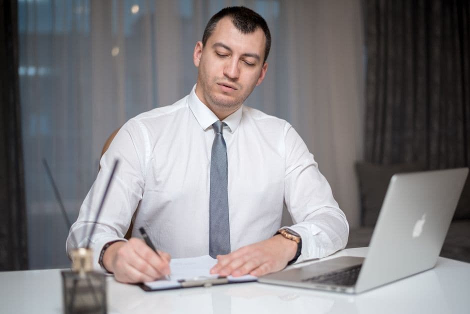 Focused businessman in office writing notes on paper with laptop at a white desk.