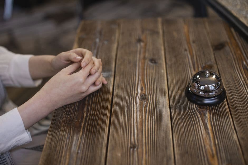 Hands folded near a call bell on a rustic wooden table, suggesting a reception or service setting.