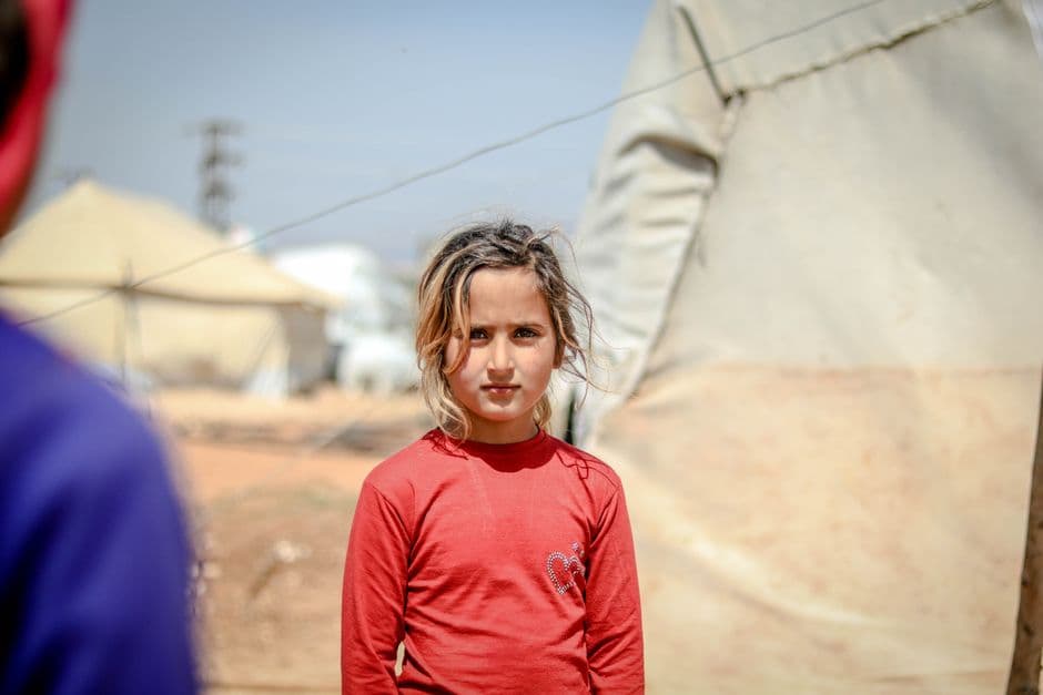 A young girl in a red shirt stands in a refugee camp in Idlib, Syria, surrounded by tents on a sunny day.