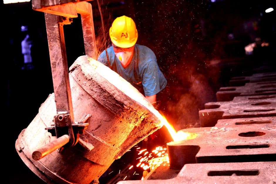 A factory worker wearing a helmet pours molten metal into molds, showcasing industrial metal casting.