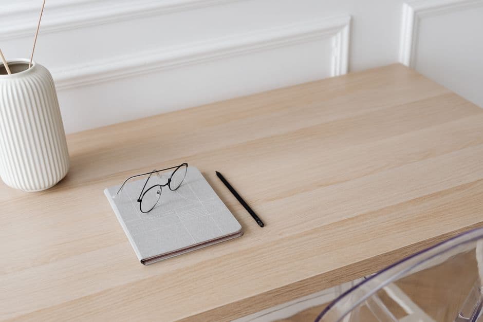 A minimalist workspace featuring a notebook, eyeglasses, pen, and a decorative vase on a wooden desk.