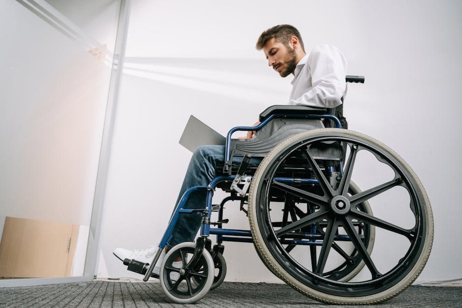 A man in a wheelchair concentrates on work using a laptop in an office setting.