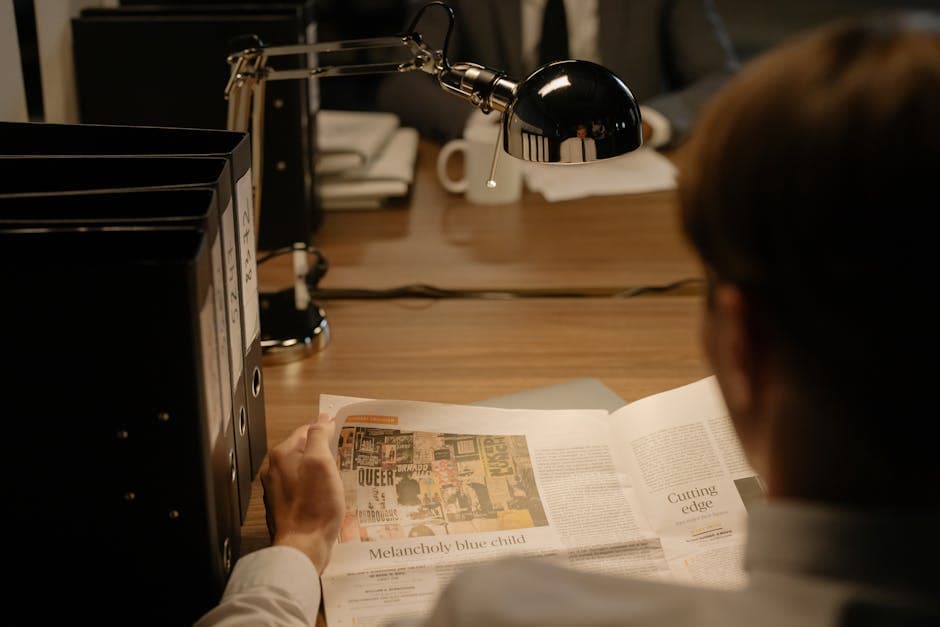 A person reads a newspaper at a desk with a lamp, suggesting investigation work.