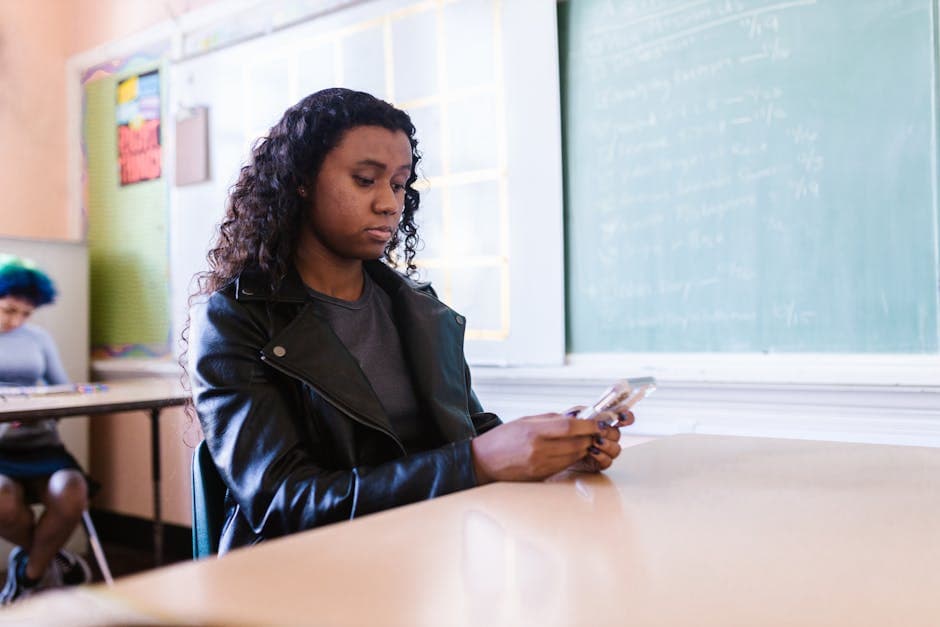 African American woman in a classroom browsing her smartphone wearing a leather jacket.