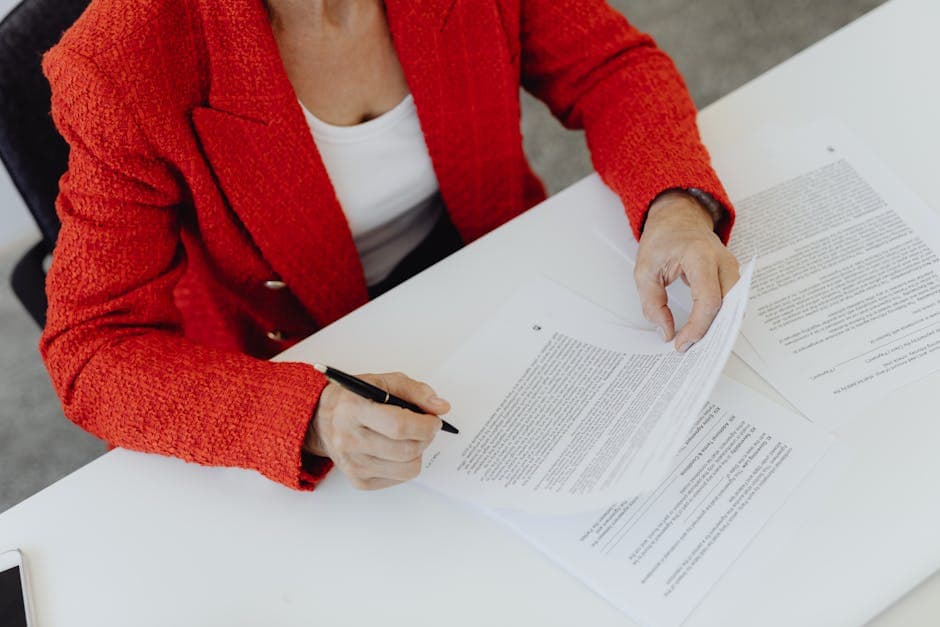 Woman in red blazer signs paperwork at a desk, emphasizing business responsibility.