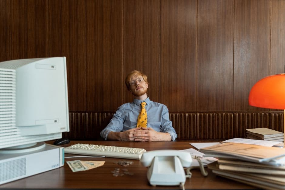 A man in a retro-style office sits at a desk with vintage technology, featuring a yellow tie and eyeglasses.