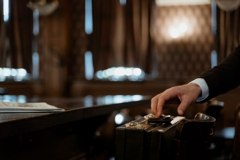 A close-up shot captures a hand holding a black briefcase in a dimly lit elegant room with a blurred background.
