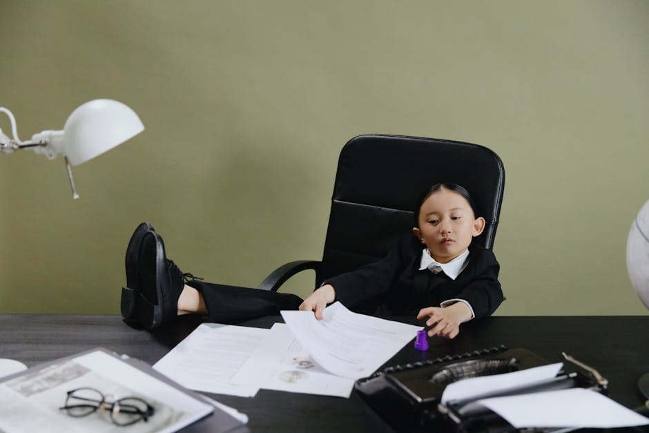 A young girl in a suit sits at a desk with papers, portraying a business theme.