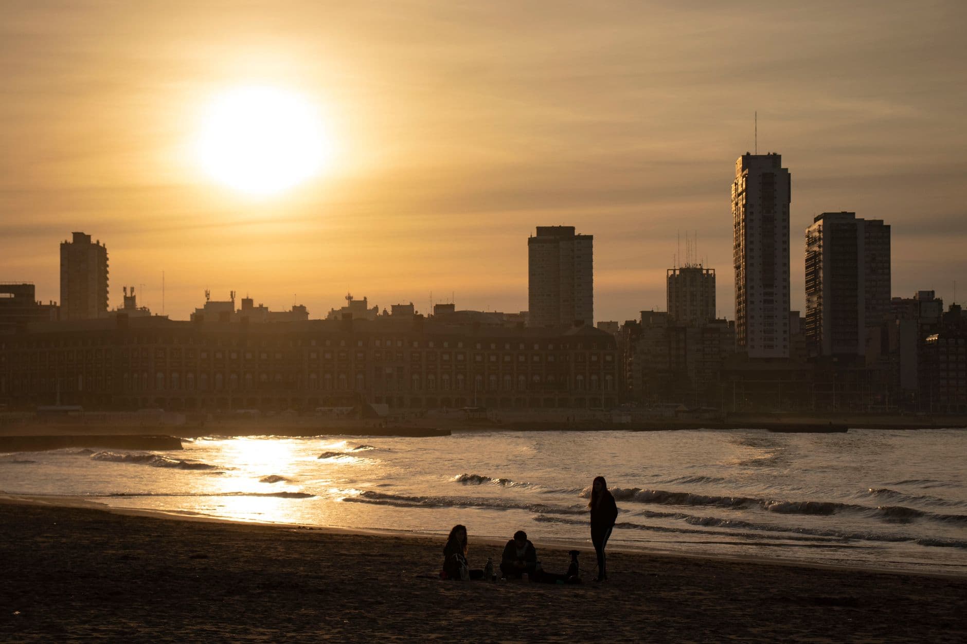 Silhouetted figures on Mar del Plata beach at sunset with city skyline in view.