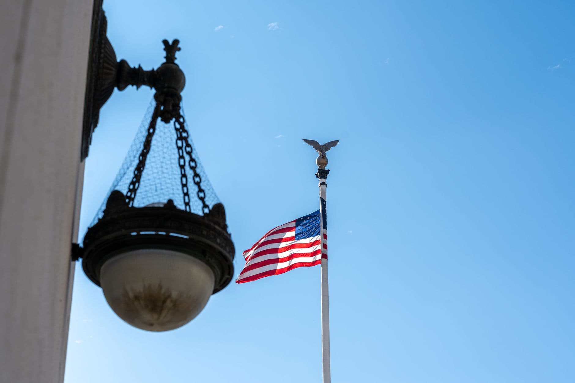 A low angle view of an American flag on a flagpole with a decorative lamp under a clear blue sky.