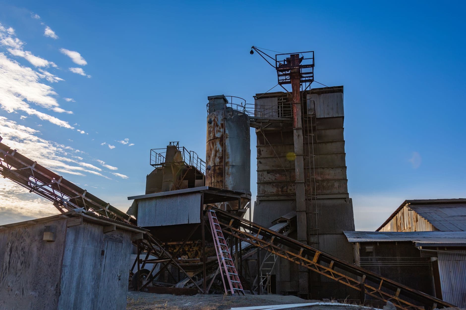 Rusty industrial facility with silos and machinery set against a blue twilight sky, depicting decay and abandonment.