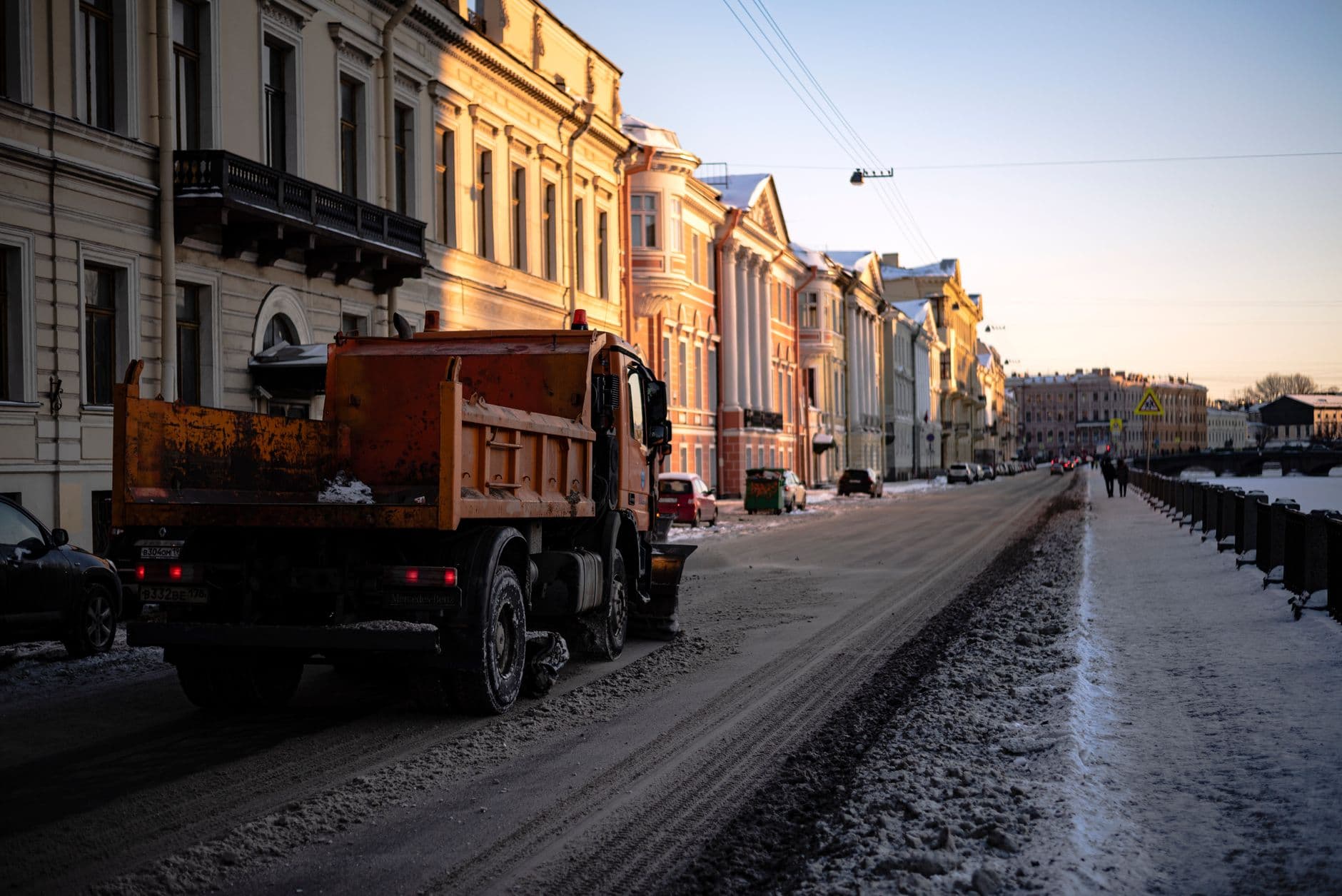 An orange truck drives down a snowy city street during sunset in winter.