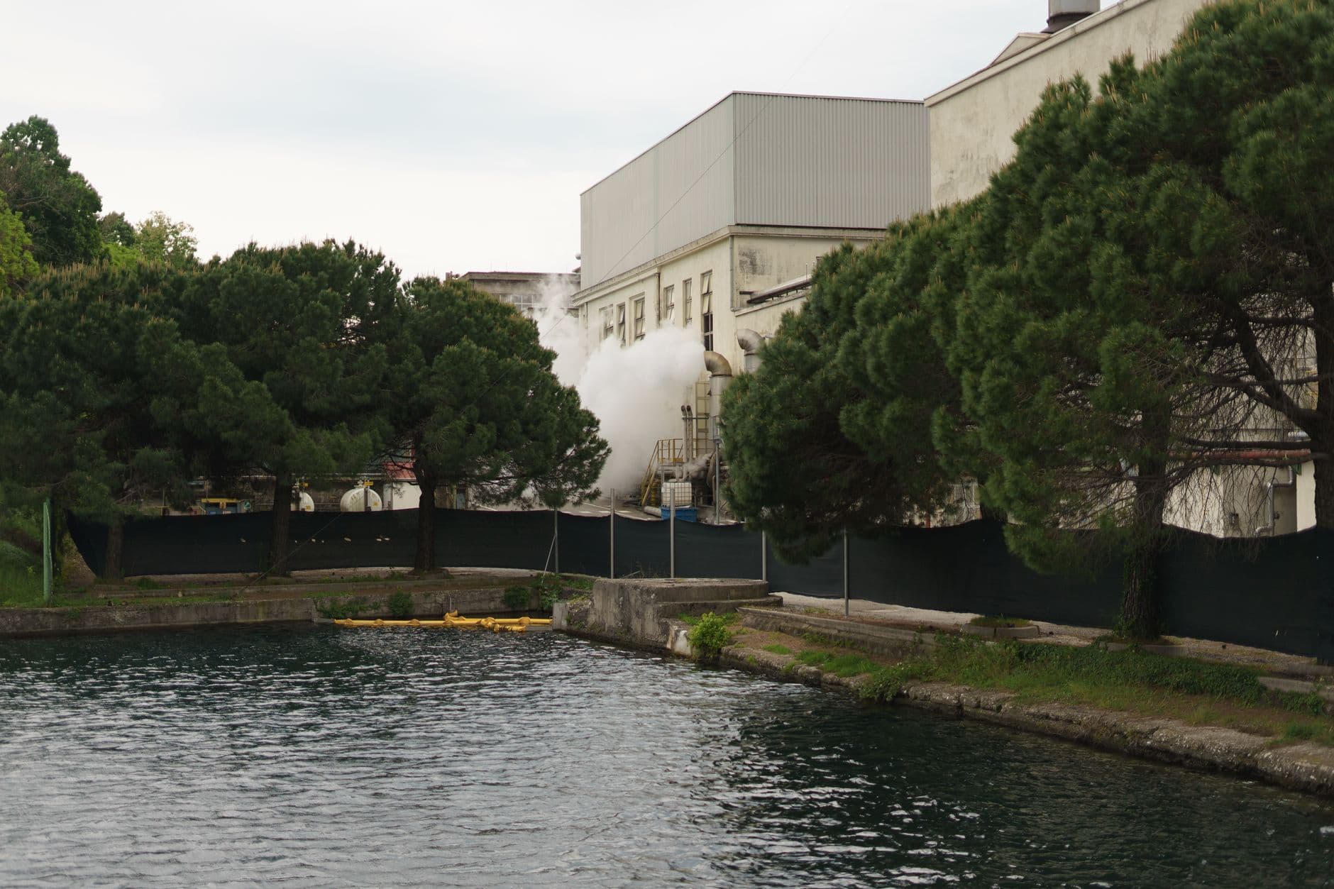 View of an industrial building with steam by a lakeside, surrounded by trees in Toscolano Maderno, Italy.