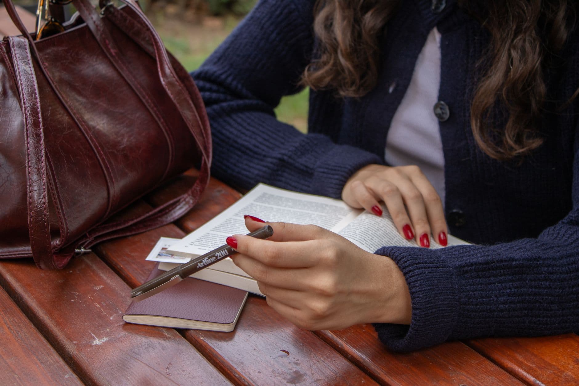 A woman with red nails reading a book and taking notes at an outdoor cafe table.