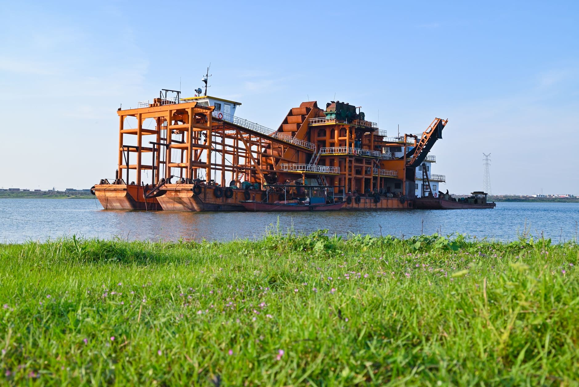 A large orange dredger barge floating on a calm lake with green grass in the foreground.