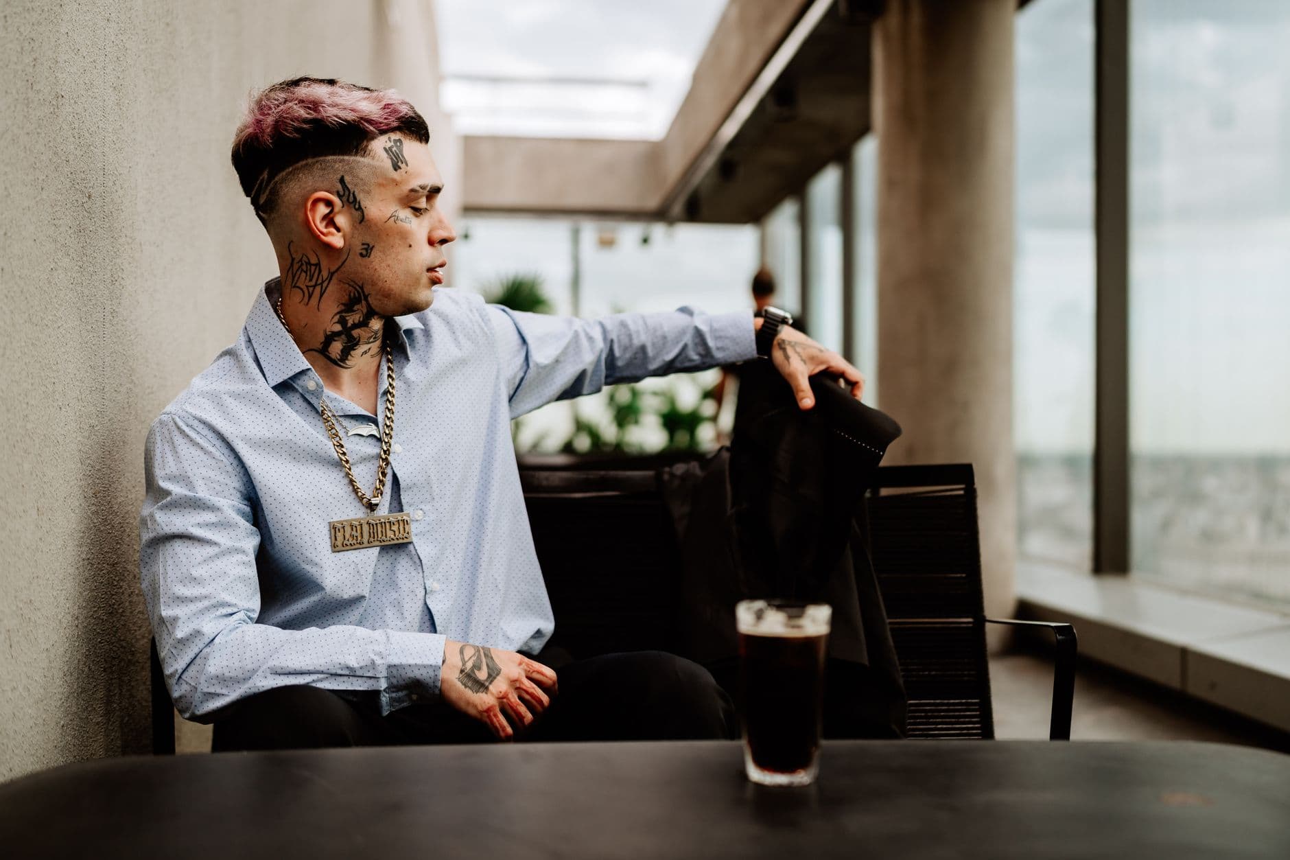 Tattooed man sitting at a table, wearing a chain necklace in a modern urban environment.