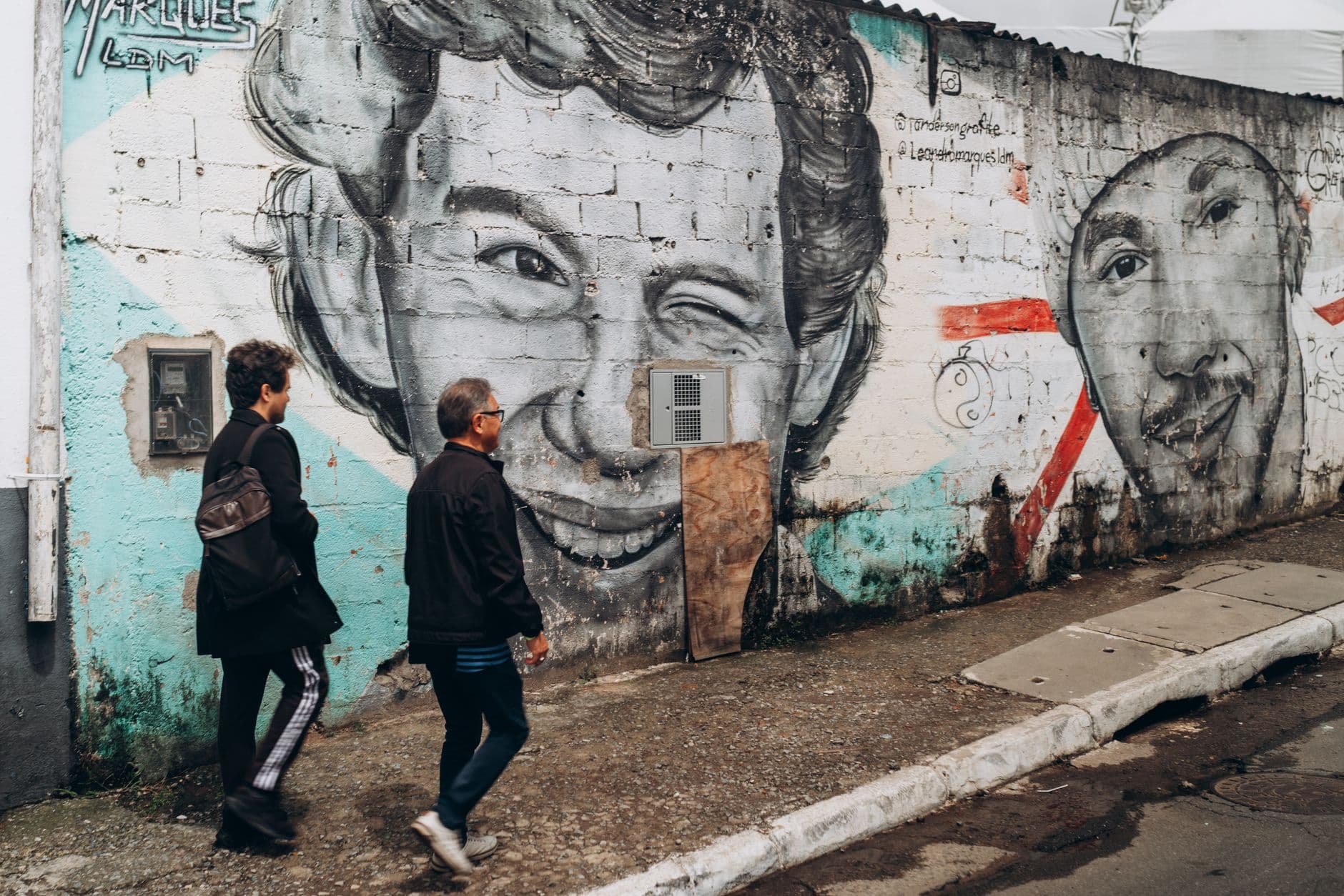 Street art mural with iconic portraits on a wall in São Paulo, Brazil. Two people walk by.