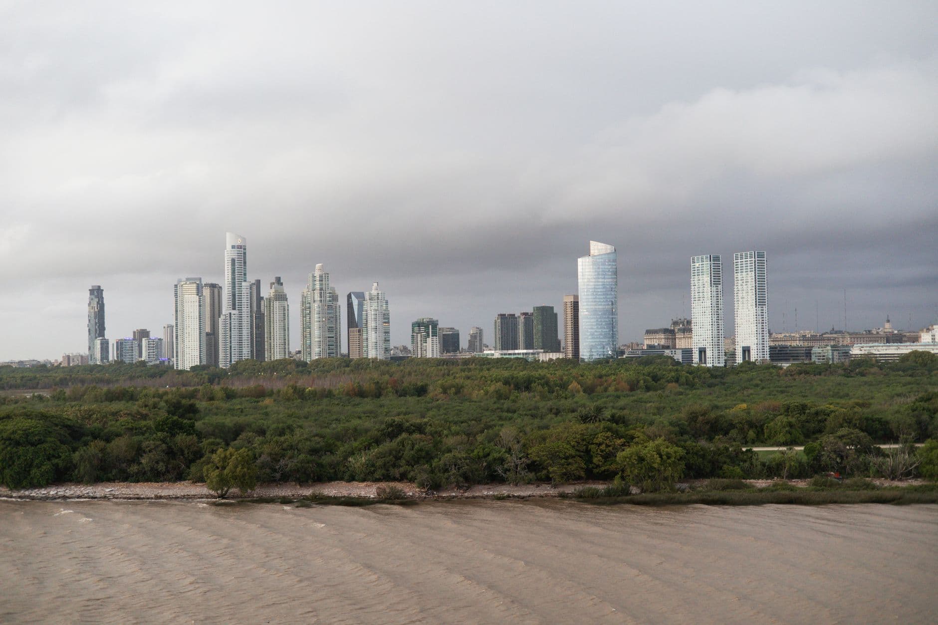 Panoramic view of Buenos Aires skyline with lush green forest and Rio de la Plata in the foreground.