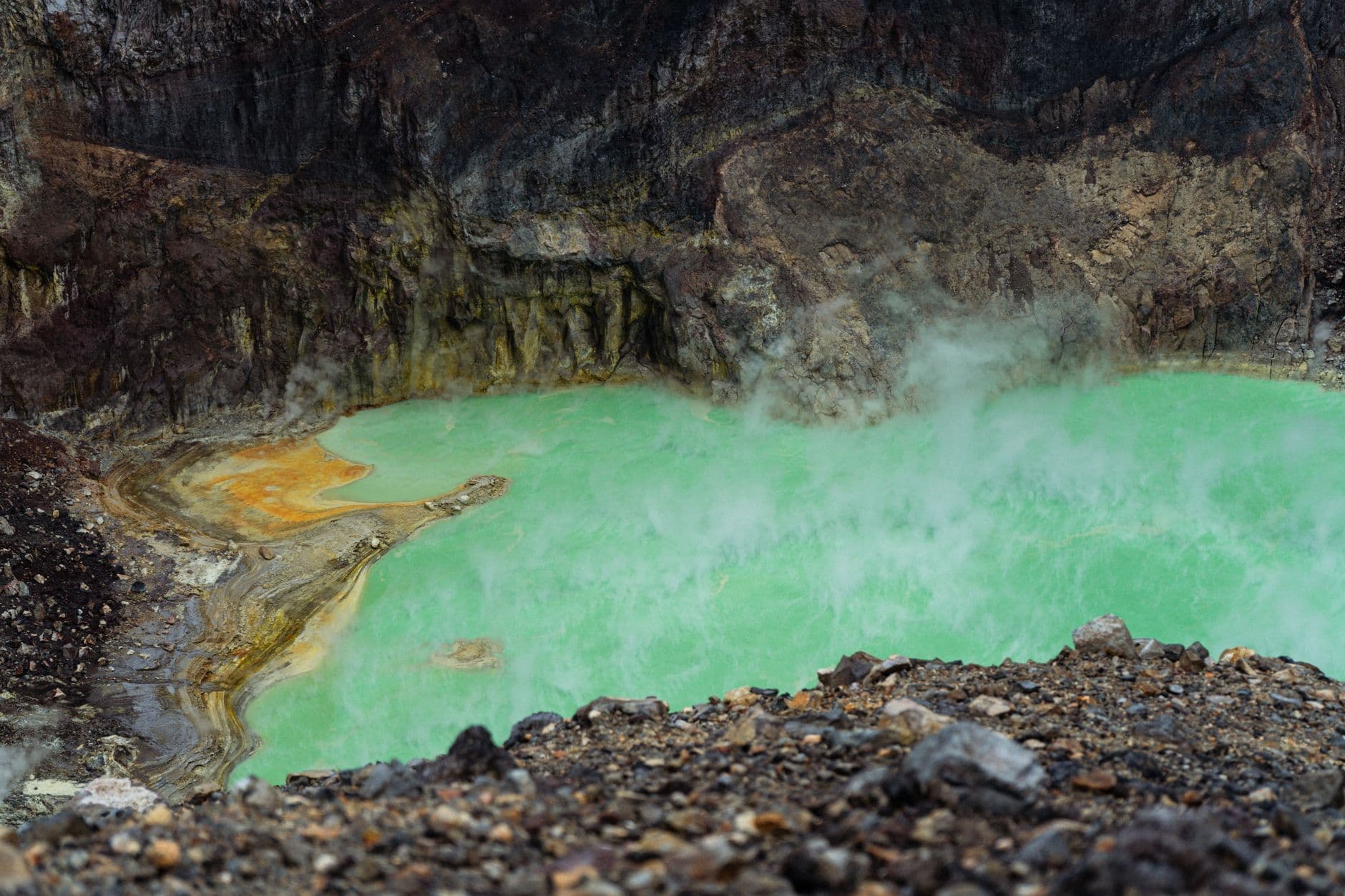 A steaming green acidic lake in a volcanic crater with vibrant color contrast against rocky terrain.
