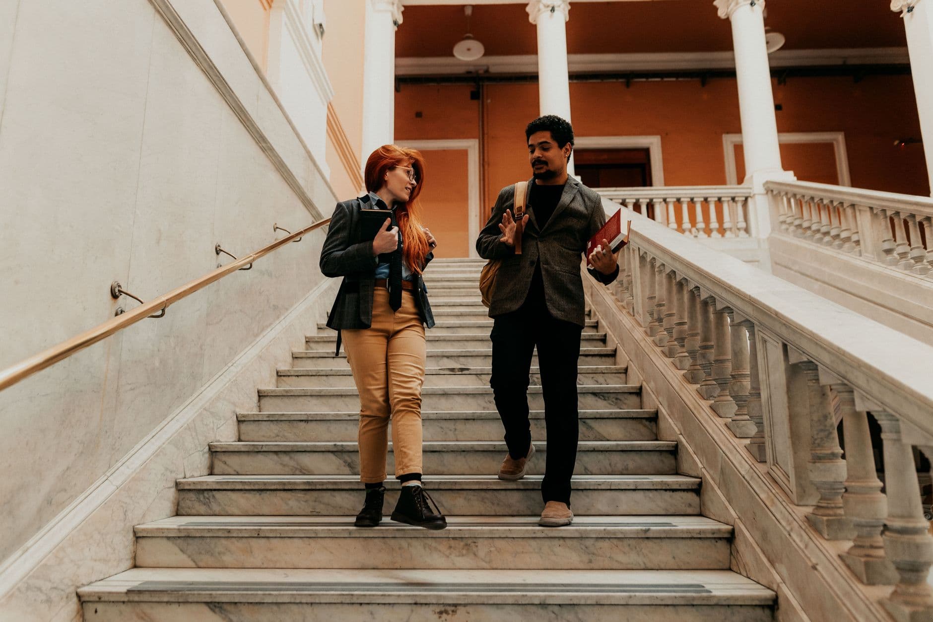 Two university students engaged in conversation while descending marble stairs inside a classic academic building.