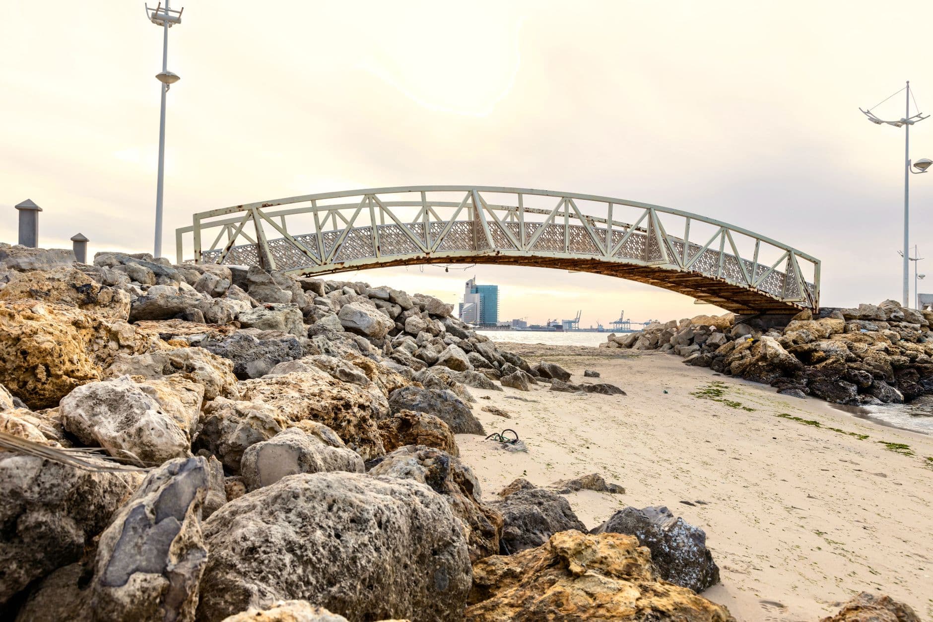 Scenic view of a rustic metal bridge over rocks on a sandy beach during sunset.