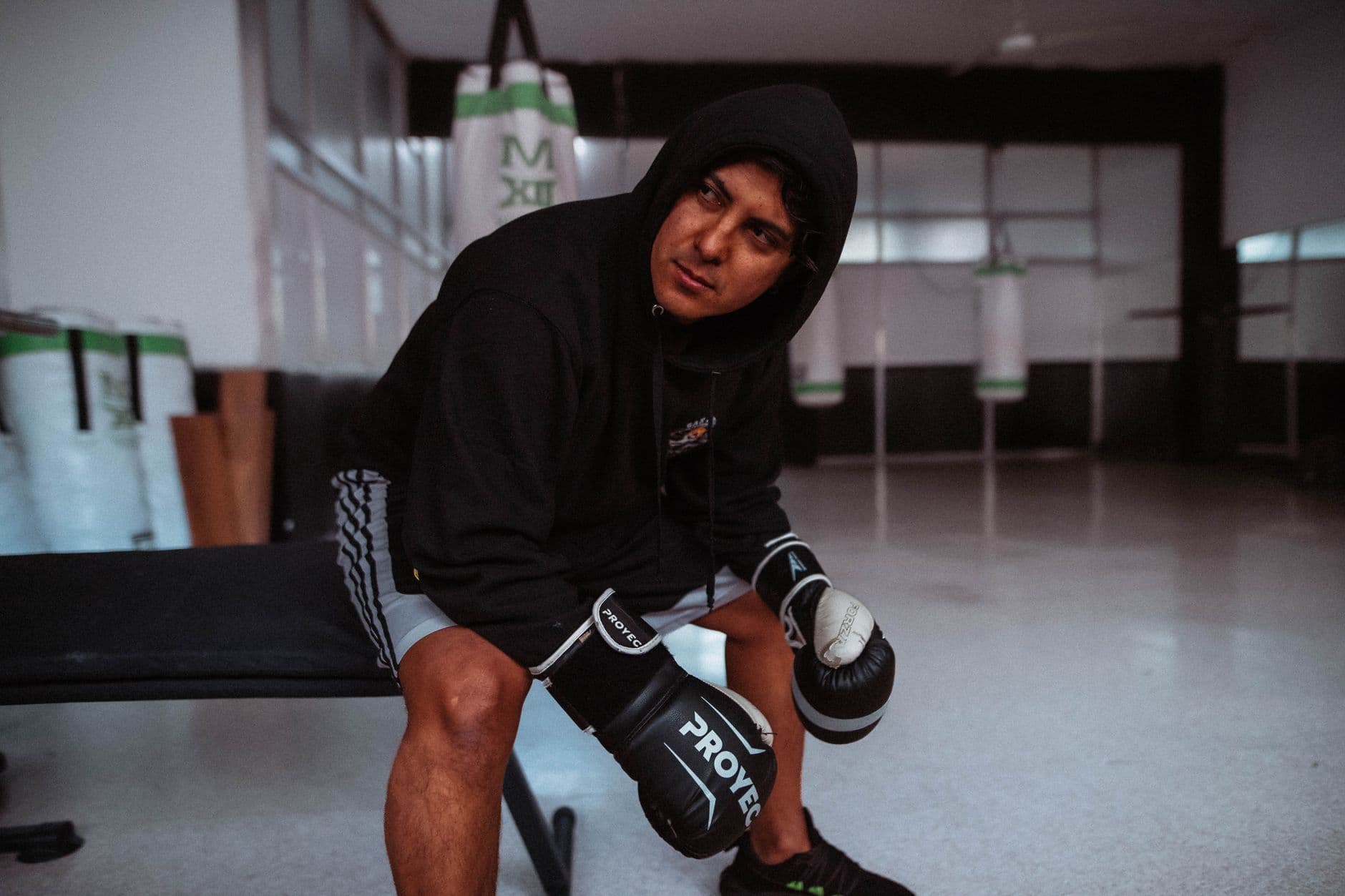 A focused boxer in a gym sitting on a bench with punching bags in the background.