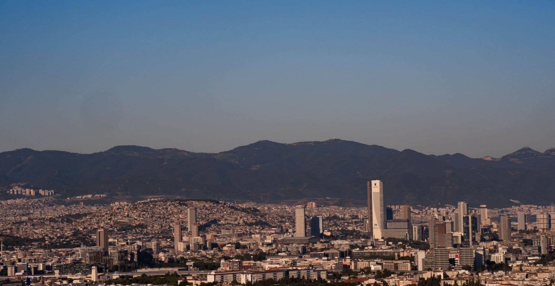 A panoramic view of İzmir city skyline with distant hills under a clear blue sky.