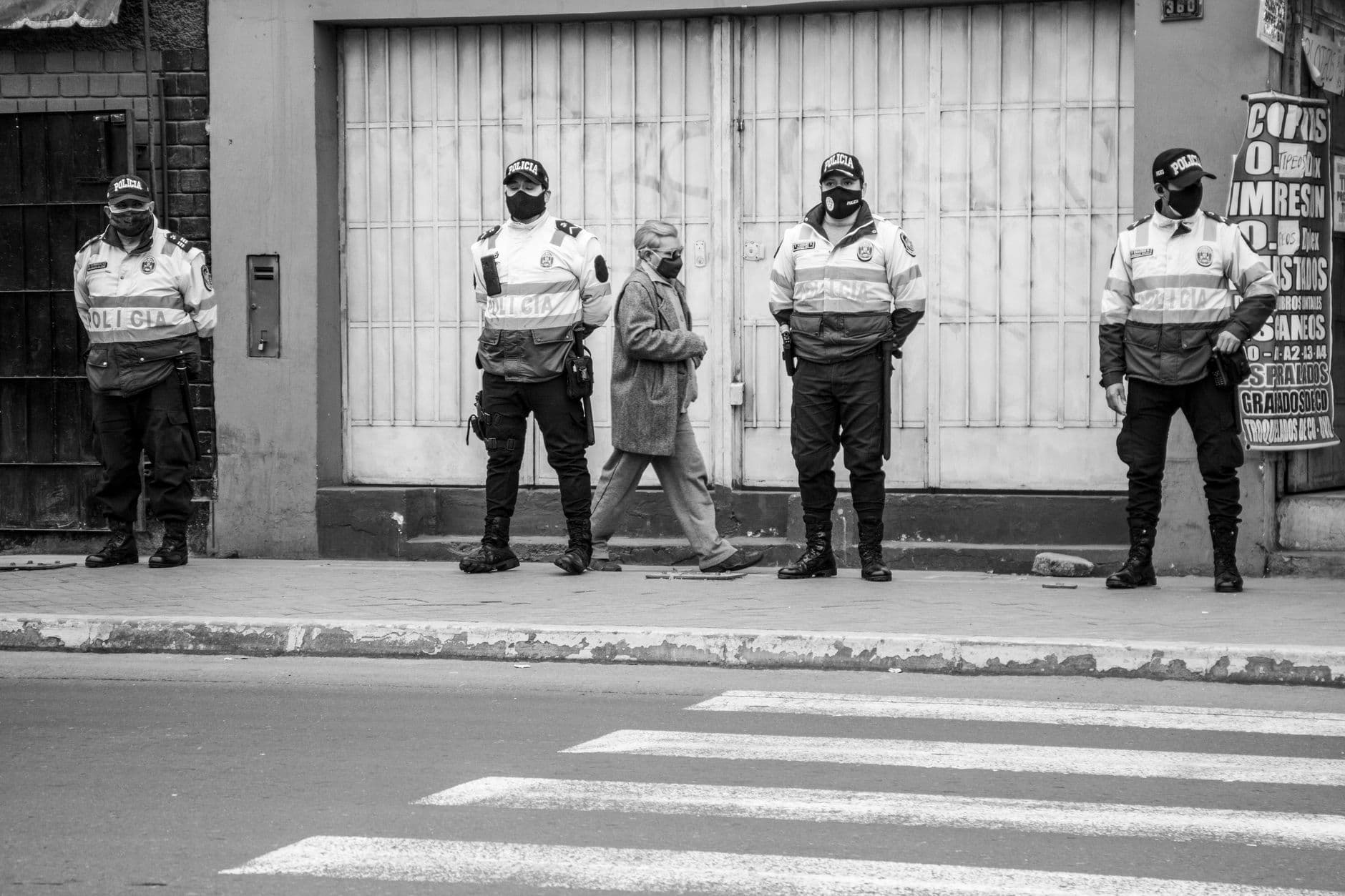 Police officers and a masked pedestrian in grayscale, maintaining social distancing on a city street.
