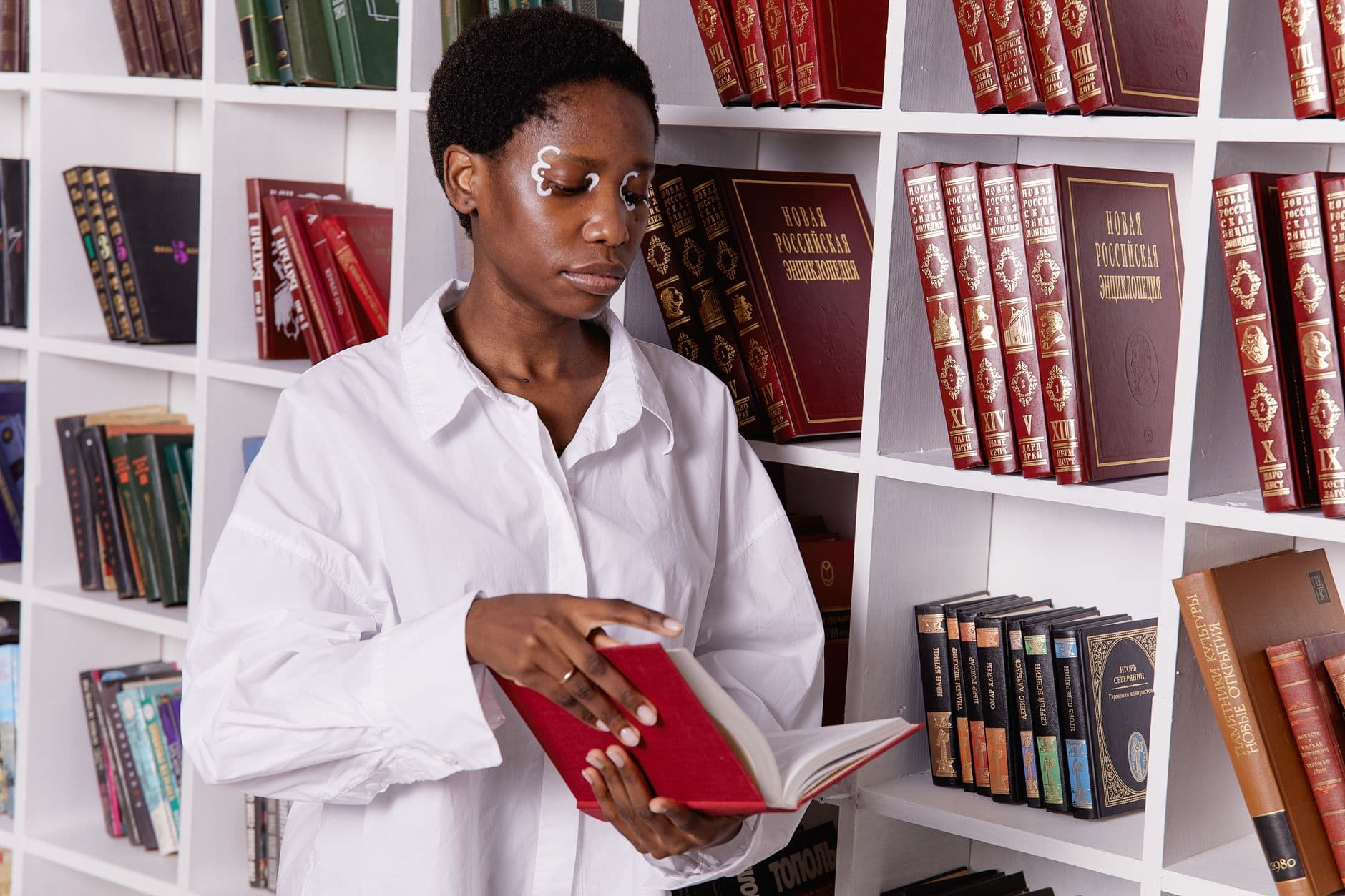 Sophisticated woman with face paint reading a book in a contemporary library setting.